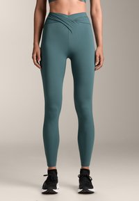 Tirkizne tajice visokog struka s uvijenim prednjim dijelom, napravljene od glatkog, rastezljivog materijala. U kombinaciji s crnim sportskim cipelama.