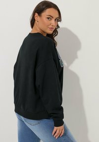 Sudadera negra de gran tamaño con puños y dobladillo acanalados, que presenta un diseño gráfico colorido en la parte frontal. Se lleva con jeans azul claros.