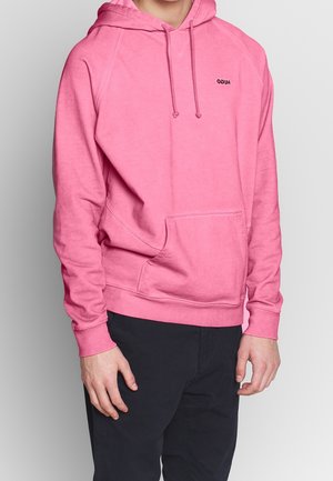 Mężczyzna w jaskraworóżowej bluzie z kapturem i kieszenią z przodu oraz czarnych spodniach, stojący na tle jednolitego szarego tła.