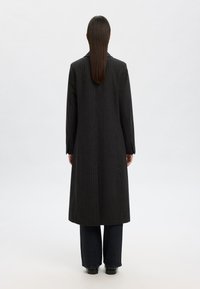 Lång svart kappa med vertikala pinstripes, skräddarsydd design, snedskurna revers och långa ärmar, bärs över mörk denimbyxor. Visas från bakåtsidan.