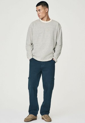 Striped long-sleeve shirt in cream and navy, paired with dark cargo pants and beige shoes. Features relaxed fit and rollable sleeves.