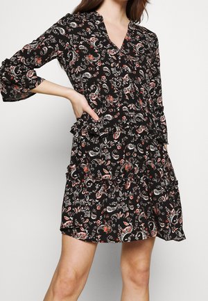 Czarna sukienka w kwiatowy wzór z rękawami do połowy przedramienia i falbankami, noszona przez kobietę pozującą z jedną ręką na biodrze.