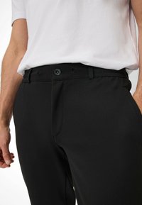 Zwarte broek heeft een gladde stof, slim fit, riemlussen en een knoopsluiting. Zakjes zijn zichtbaar aan de zijkant.