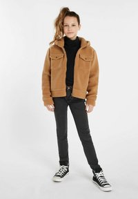 Giacca in pile marrone con zip, dotata di due tasche sul petto, abbinata a jeans neri slim e sneakers alte nere. Tessuto liscio e design con cappuccio.