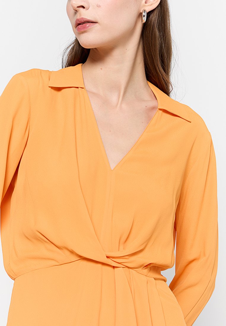 Pomarańczowa bluzka z długim rękawem, z dekoltem w kształcie litery V i kołnierzykiem. Zawiera drapowany przód i delikatną teksturę. Brak widocznych wyrazistych wzorów lub elementów metalowych.