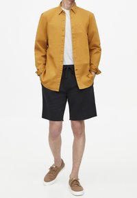 Pánský outfit obsahující hořčicově žlutou košili na knoflíky, šedé tričko, černé šortky a hnědé tenisky. Jednoduchý design s dlouhými rukávy.