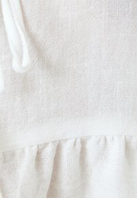 Tejido blanco ligero con un patrón texturizado y tejido. Presenta un detalle de volantes fruncidos en la parte inferior y un componente de cordón.