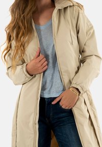 Giacca lunga khaki con cappuccio, chiusura con zip e polsini elasticizzati, indossata sopra una maglietta azzurro chiaro e jeans scuri. Tessuto morbido e resistente all'acqua.