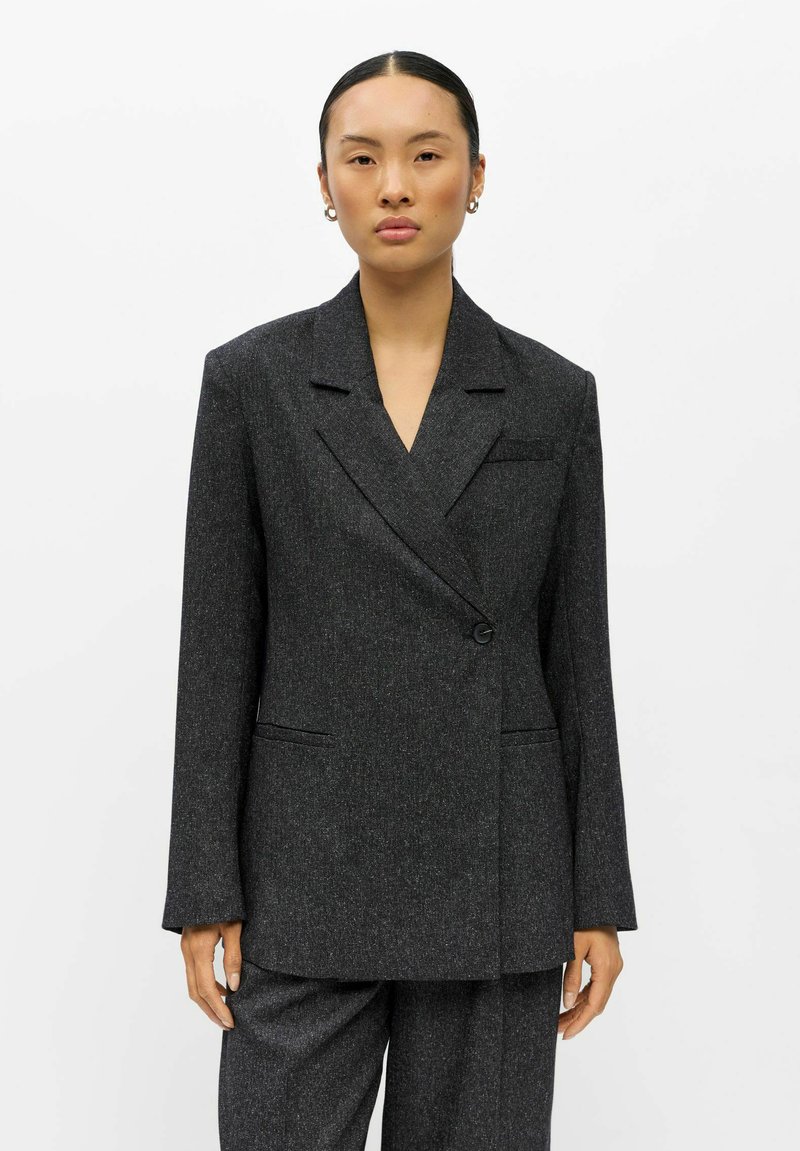 Femme aux cheveux plaqués en arrière porte un blazer oversize gris foncé et un pantalon assorti sur un fond blanc uni.