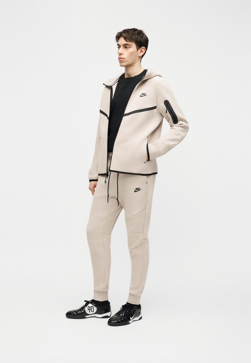 Joven de pie, vestido con chándal beige de Nike con detalles en negro, camiseta negra y zapatillas Nike negras con suelas blancas.