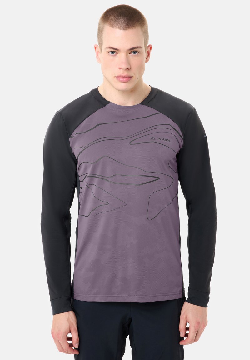 Jeune homme aux cheveux courts portant un maillot de sport Vaude à manches longues violet et noir avec des motifs de lignes abstraites, debout devant un fond clair.