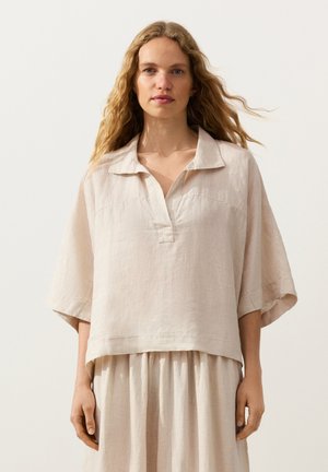 Vrouw met lang golvend blond haar, die een losse lichtbeige linnen blouse en bijpassende rok draagt, staand tegen een effen lichte achtergrond.