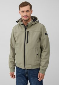 Veste softshell kaki avec capuche, fermeture éclair frontale, poche poitrine inclinée et deux poches latérales avec fermetures à bouton, associée à un jean bleu.