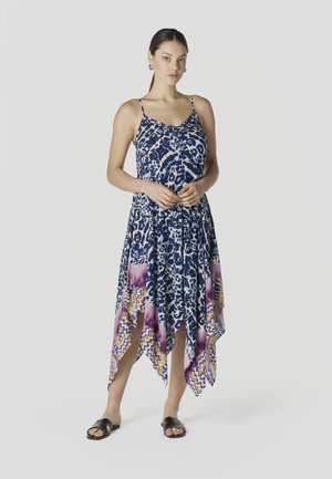 Mujer de pie con vestido sin mangas azul y blanco con estampado de animales y bajo asimétrico con patrones en púrpura y amarillo, sandalias negras tipo chancla.