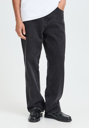 Person iført løstsiddende sorte jeans, sorte skinnende pæne sko og en hvid skjorte med den ene hånd i lommen mod en ensfarvet baggrund.