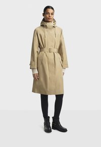 Beige waterdichte trenchcoat met een tailleceintuur, capuchon en grote zijzakken. Gecombineerd met zwarte enkellaarsjes en donkere leggings.