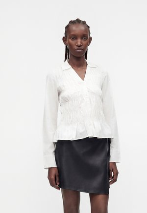 Femme aux cheveux tressés portant un chemisier blanc à manches longues et une jupe en cuir noire asymétrique, debout sur un fond uni.