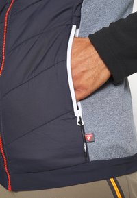 Modrý a šedý vest, který je vyroben z prošívaného námořnického materiálu a má červené akcenty. Obsahuje kapsu na zip a fleecovou podšívku.