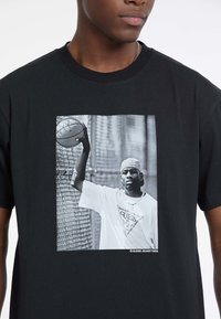 T-shirt noire arborant un graphique noir et blanc marquant d'un homme tenant un ballon de basket, portant un t-shirt avec logo blanc et une casquette.