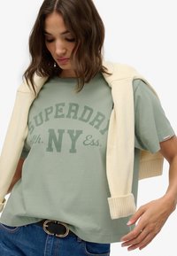 Camiseta de algodón verde claro con la impresión en verde oscuro de "SUPERDRY NY Ess." Suéter crema caído sobre los hombros y cinturón negro con hebilla dorada.
