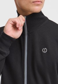 Schwarze Zip-Jacke mit hohem Kragen, gerippten Bündchen und grauem Reißverschluss. Verfügt über ein kleines Logo auf der Brust. Weicher Stoff.