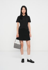 Sort polo t-shirt med knapper, parret med en sort nederdel. Modellen holder en mønstret håndtaske, iført hvide sokker og sorte loafers.