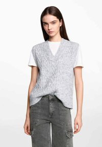 Grå stickad väst med V-ringning, med en kabelstickad mönster. Bärs över en vit t-shirt och är kombinerad med grå cargobyxor.