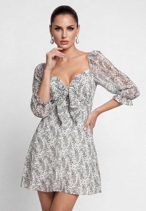 Frau mit glattem Haar, die Perlenohrringe mit Anhängern und ein weißes Kleid mit schwarzem Sprenkelmuster, Puffärmeln und einer Schleifendetail am Dekolleté trägt.