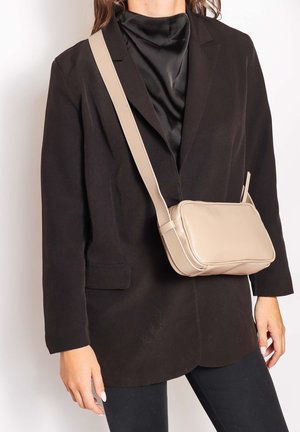 Frau trägt schwarzen Blazer und Satin-Top, hält beigefarbene, rechteckige Umhängetasche mit breitem Riemen.