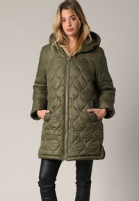 Olijfgroen gewatteerde jas met een rits, capuchon en twee zijzakken. Heeft een gestructureerd patroon en een bontvoering langs de capuchon.