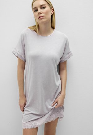 Joven con un vestido corto de manga corta color lavanda claro con ribete de encaje en las mangas y el dobladillo, de pie frente a una pared blanca lisa.