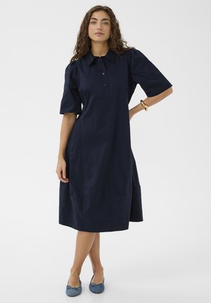 Kobieta o kręconych włosach, ubrana w granatową sukienkę do kolan, z bufkami na rękawach, kołnierzykiem i guzikami, stojąca z jedną ręką na biodrze.