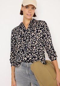 Långärmad marinblå blus med krämfärgad leopardprint, med en V-halsringning. Kombinerad med ljusgrå jeans och håller en khakifärgad kudde.