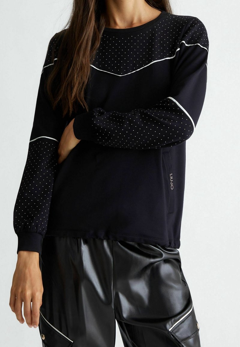 Femme portant un haut noir à manches longues avec des détails blancs à pois et un pantalon noir brillant avec des poches zippées, debout devant un fond uni.