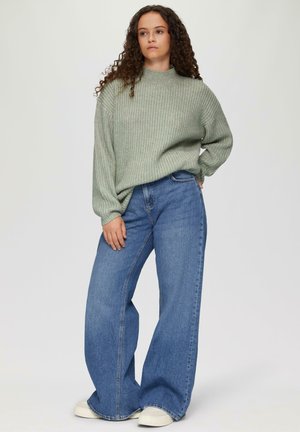 Hellgrüner gerippter Strickpullover mit hohem Kragen, kombiniert mit weiten blauen Jeans und weißen Sneakern.