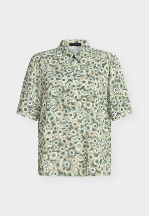 Femme aux cheveux blonds courts portant une chemise verte à fleurs à manches courtes, un pantalon blanc à jambes larges, des chaussures noires, tenant un sac à main noir.