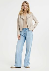 Giacca da biker in pelle beige chiaro con chiusura a zip, abbinata a jeans a gamba larga blu chiaro e sneakers beige. Texture liscia e design aderente.