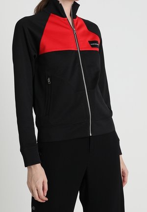 Persona vistiendo una chaqueta con cremallera negra y roja, con un pequeño bolsillo en el pecho y mangas largas, combinada con pantalones negros sobre un fondo liso.