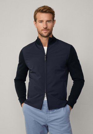 Veste navy avec fermeture éclair à l'avant, en tissu mélangé : partie supérieure lisse et manches texturées. Comprend un col montant et une coupe ajustée. Pantalons bleu clair.