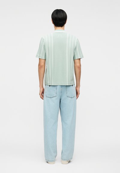 Άνδρας που φοράει ανοιχτό πράσινο κοντομάνικο πουκάμισο με κάθετες ρίγες και ανοιχτό μπλε φαρδιά τζιν, στέκεται με την πλάτη στραμμένη σε απλό φόντο.