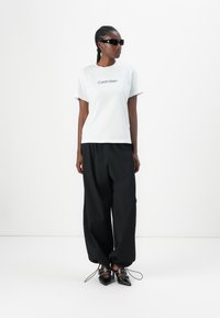 Camiseta blanca de algodón con el logo "Calvin Klein", combinada con pantalones negros sueltos y zapatos negros de punta con detalles de correa.