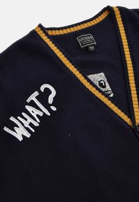 Cardigan tricoté bleu marine avec bordure jaune, texte blanc brodé "WHAT?" sur la poitrine, et étiquette de la marque Guess à l'intérieur du col.