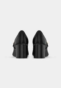 Paire de chaussures en cuir noir à talon bloc vues de dos avec un détail de boucle dorée sur les côtés sur fond gris.