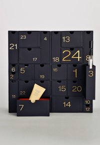 Calendario dell'Avvento nero con numeri dorati sui cassetti. Contiene vari scomparti. Uno scomparto mostra un tubo giallo e un altro contiene uno spray.
