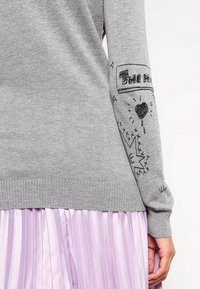 Manche de pull gris avec des griffonnages noirs incluant un cœur, des étoiles et du texte, associée à une jupe plissée violet clair.