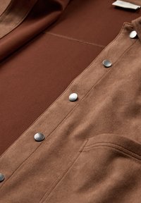 Brunt mockaskinn med en slät yta, prydd med silverfärgade tryckknappar och en framficka, mot en enfärgad brun bakgrund.