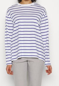 Persona che indossa una camicia bianca a maniche lunghe con sottili strisce orizzontali blu e pantaloni grigio chiaro, in piedi su uno sfondo semplice.