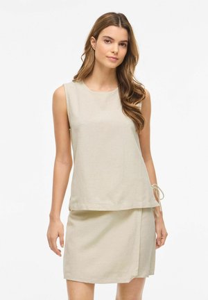 Młoda kobieta z długimi falującymi włosami, ubrana w beżowy bezrękawnik i dopasowaną spódnicę do kolan, stojąca na jednolitym białym tle.