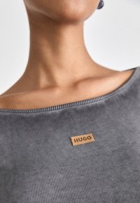 Siva rebrasta majica z širokim izrezom in svetlo rjavim usnjenim oznako z vdelanim napisom "HUGO" na sprednji strani. Mehak videz, minimalističen dizajn.
