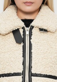 Närbild av en person som bär en krämfärgad sherpa-jacka med svarta läderdetaljer, en dragkedja fram och spännedetaljer på kragen.